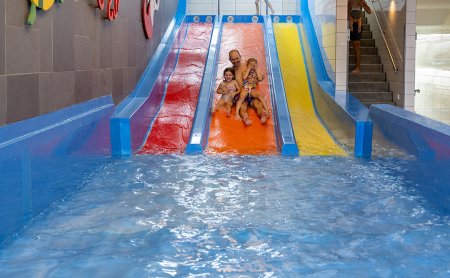 Vater mit zwei Kindern rutscht eine orange Wasserrutsche in einem Hallenbad hinunter