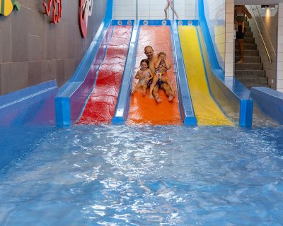 Vater mit zwei Kindern rutscht eine orange Wasserrutsche in einem Hallenbad hinunter