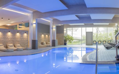 Innenaufnahme eines modernen Schwimmbads mit blau beleuchtetem Wasser, Liegestühlen und Sitzkissen