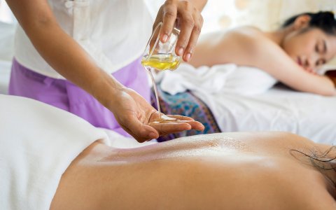 Nahaufnahme einer Hand, die Massageöl auf den Rücken einer liegenden Person gießt, mit einer weiteren Person im Hintergrund