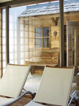 Drei weiße Liegestühle vor einer Glaswand mit Blick auf eine Holzhütte im Innenbereich