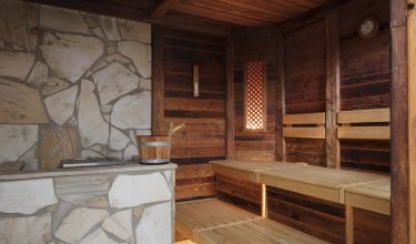 Innenraum einer Sauna mit Holzbänken, Steinofen und Holzeimer auf dem Ofen