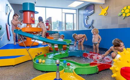 Kinder spielen an einem bunten Wasserspiel mit Rutschen und Bällen in einem hellen Raum mit Fenster