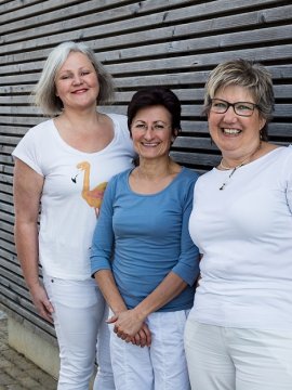 Drei lächelnde Frauen stehen nebeneinander vor einer Holzlattenwand im Freien.