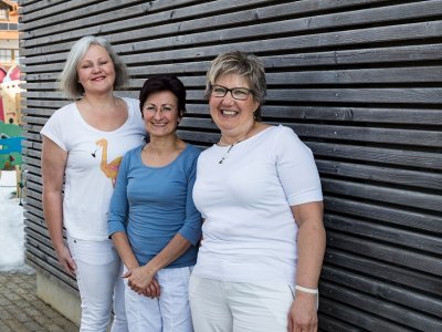 Drei lächelnde Frauen stehen nebeneinander vor einer Holzlattenwand im Freien.