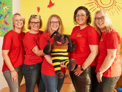 Fünf Frauen in roten T-Shirts halten ein großes Plüschtier in einem gelben Raum mit Schmetterlings- und Sonnenwanddekoration