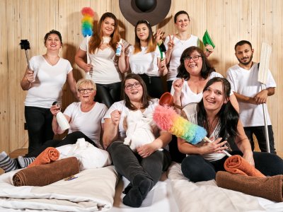 Gruppe von Menschen in weißen T-Shirts mit Reinigungsutensilien vor Holzwand, lächelnd