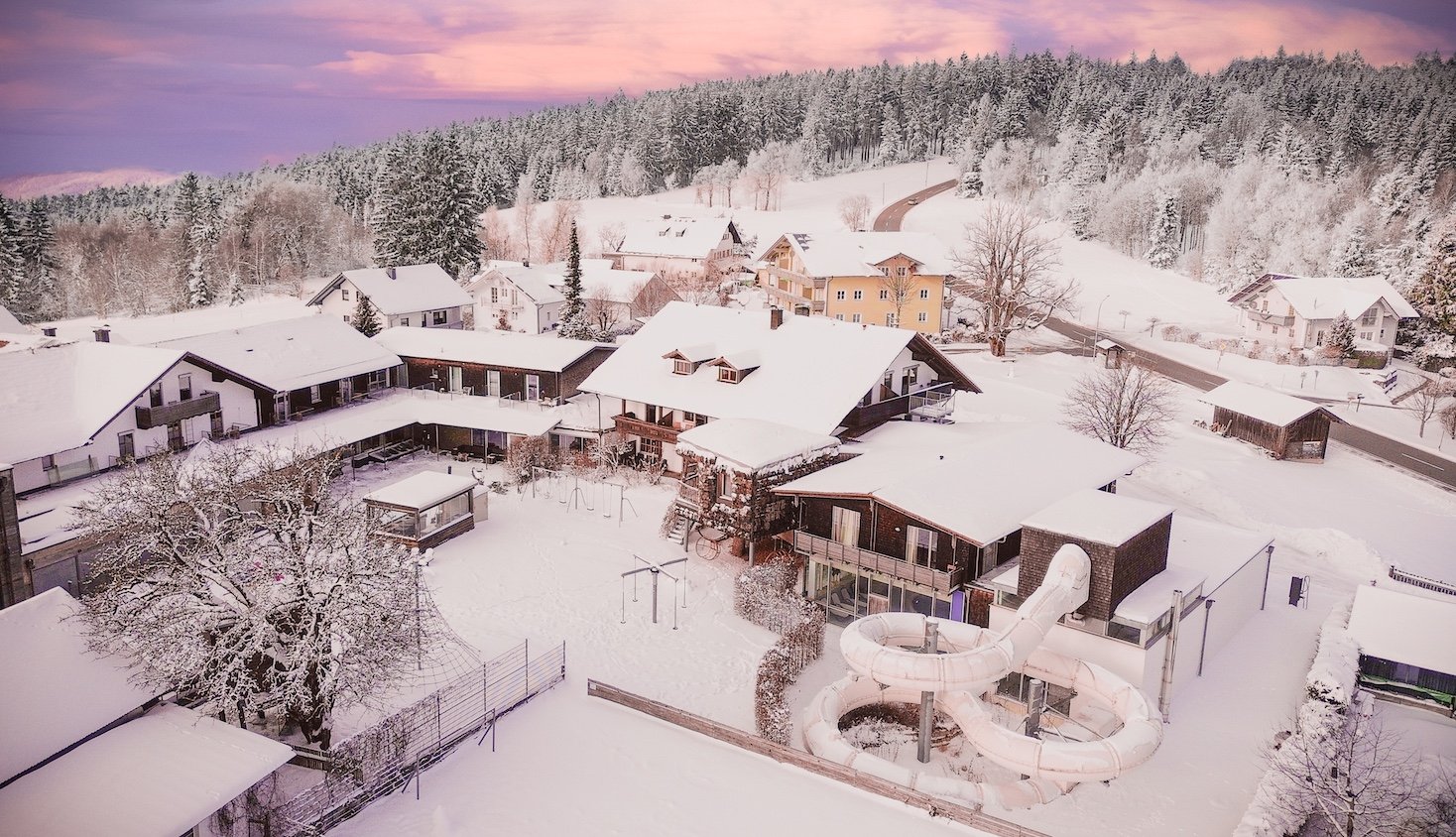 Verschneites Dorf mit Häusern und einer Wasserrutsche vor einem Wald bei Sonnenuntergang