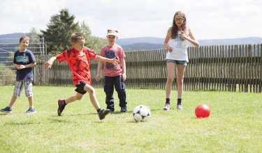 Kinder spielen Fußball auf einer Wiese vor einem Holzzaun bei sonnigem Wetter