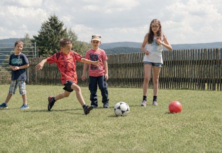 Kinder spielen Fußball auf einer Wiese vor einem Holzzaun und Bäumen im Hintergrund