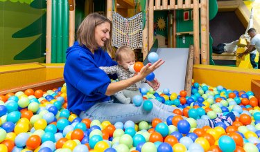 Frau und Baby sitzen im Bällebad eines Spielplatzes, umgeben von bunten Bällen