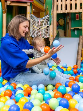 Frau und Baby sitzen im Bällebad eines Spielplatzes, umgeben von bunten Bällen