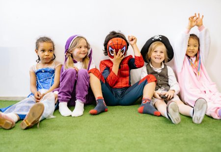 Fünf Kinder in Kostümen sitzen nebeneinander auf grünem Teppich vor weißer Wand