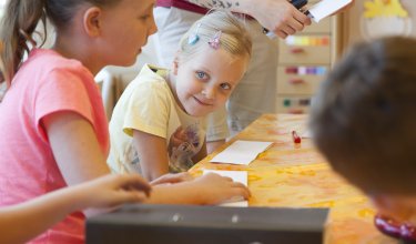 Kinder sitzen an einem Tisch mit buntem Tischtuch, ein Mädchen schaut lächelnd zur Seite
