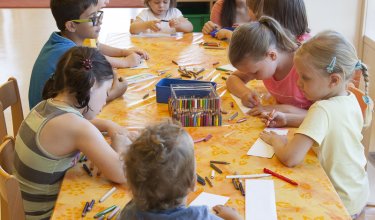 Kinder sitzen an einem Tisch mit orangener Tischdecke und malen mit Buntstiften in einem hellen Raum