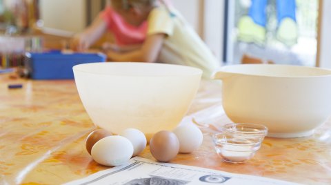 Zwei weiße Schüsseln, Eier und kleine Schalen mit Zutaten auf einem Tisch, im Hintergrund unscharfe Kinder
