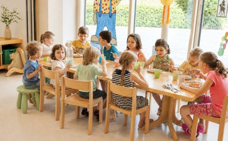 Kinder sitzen an einem Holztisch und essen gemeinsam in einem hellen Raum mit großen Fenstern