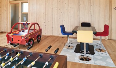 Spielzimmer mit Tischkicker im Vordergrund, rotem Airhockey-Auto links und bunten Stühlen um einen Tisch