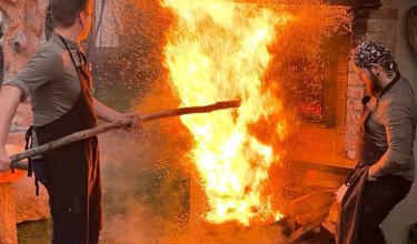 Zwei Männer in Schürzen stehen vor einem großen Feuer mit Funken auf gepflastertem Boden im Freien