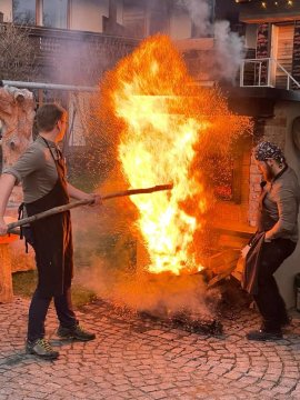 Zwei Männer in Schürzen stehen vor einem großen Feuer mit Funken auf gepflastertem Boden im Freien