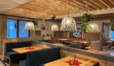 Gemütliches Restaurant mit Holztischen, grünen Sitzbänken, hängenden Pflanzen und Lampen