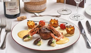 Gourmetgericht mit Garnelen, Fleisch, Kartoffeln und Sauce auf weißem Teller, Weinflasche im Hintergrund