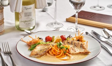 Gebratenes Fischfilet mit Gemüse und Soße auf weißem Teller, Weinflasche und Gläser im Hintergrund
