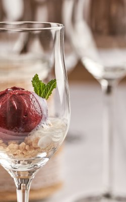 Nahaufnahme eines Weinglases mit rotem Sorbet, Minzblatt und Keksbröseln, unscharfe Gläser im Hintergrund