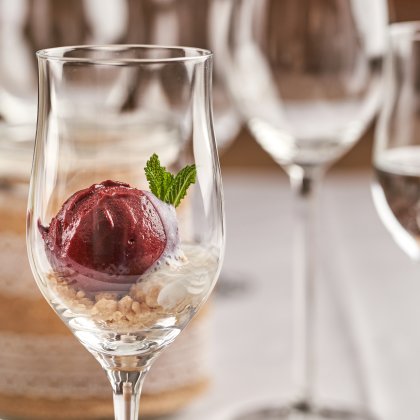 Nahaufnahme eines Weinglases mit rotem Sorbet, Minzblatt und Keksbröseln, unscharfe Gläser im Hintergrund
