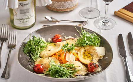 Teller mit Ravioli, Rucola, Kirschtomaten und Parmesan, daneben Besteck und Weinflasche