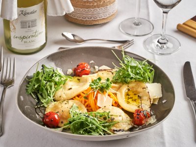 Teller mit Ravioli, Rucola, Kirschtomaten und Parmesan, daneben Besteck und Weinflasche