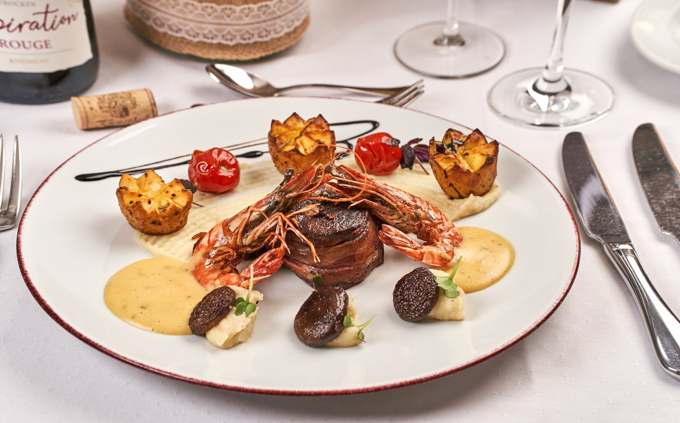 Gourmetgericht mit Garnelen, Steak, Kartoffeln und Sauce auf weißem Teller, Besteck und Weinglas