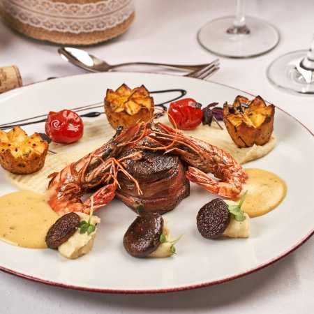 Gourmetgericht mit Garnelen, Steak, Kartoffeln und Sauce auf weißem Teller, Besteck und Weinglas