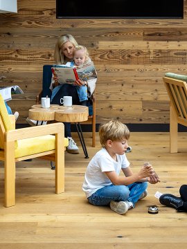 Kinder sitzen auf dem Boden und spielen Karten, während Erwachsene auf Stühlen im Hintergrund Bücher lesen