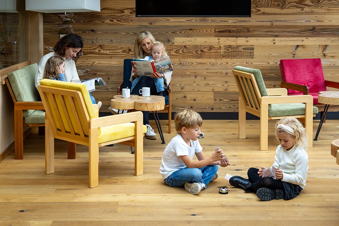 Kinder sitzen auf dem Boden und spielen Karten, während Erwachsene auf Stühlen im Hintergrund Bücher lesen