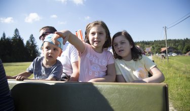 Kinder sitzen auf einer offenen Fahrt im Freien, ein Mädchen zeigt nach vorne, ländliche Umgebung im Hintergrund