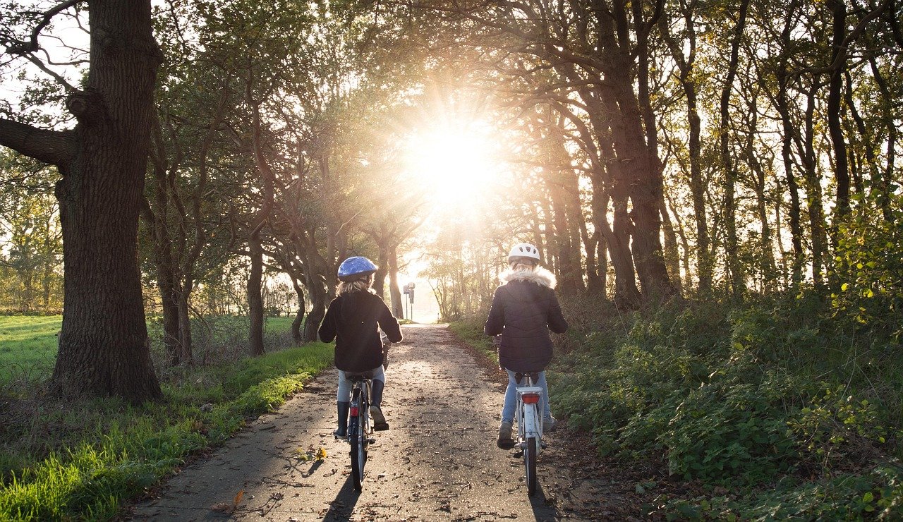 Zwei Kinder fahren mit Helmen auf einem von Bäumen gesäumten Weg in die Sonne