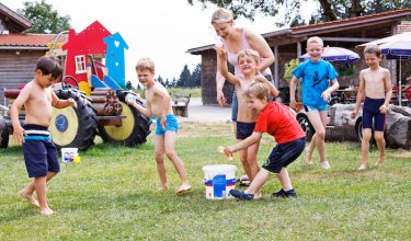 Kinder spielen mit Wasserbällen auf einer Wiese, eine Frau beobachtet sie lächelnd