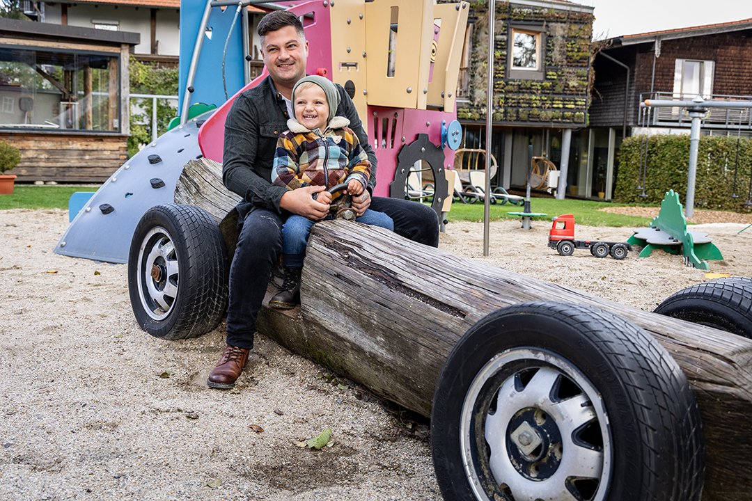 Vater und Kind sitzen lachend auf einem Spielplatz-Holzauto mit Reifen und buntem Klettergerüst im Hintergrund