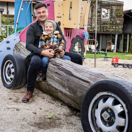 Vater und Kind sitzen lachend auf einem Spielplatz-Holzauto mit Reifen und buntem Klettergerüst im Hintergrund