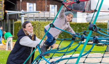 Frau unterstützt Kind beim Klettern auf einem blauen Kletternetz auf einem Spielplatz vor einem Haus