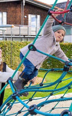 Frau unterstützt Kind beim Klettern auf einem blauen Kletternetz auf einem Spielplatz vor einem Haus