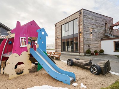 Bunter Kinderspielplatz mit Rutsche und Holzauto vor modernem Holzhaus an bewölktem Tag