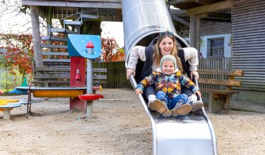 Frau und Kind rutschen zusammen eine Metallrutsche auf einem Spielplatz im Freien hinunter