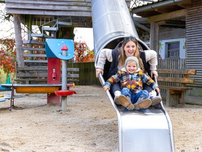 Frau und Kind rutschen zusammen eine Metallrutsche auf einem Spielplatz im Freien hinunter