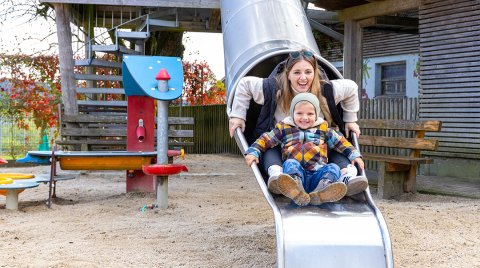 Frau und Kind rutschen zusammen eine Metallrutsche auf einem Spielplatz im Freien hinunter
