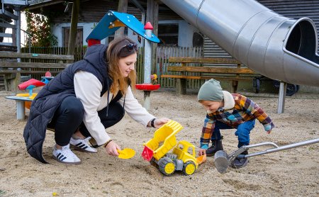 Frau und Kind spielen mit gelbem Spielzeugbagger im Sandkasten eines Spielplatzes neben einer Rutsche