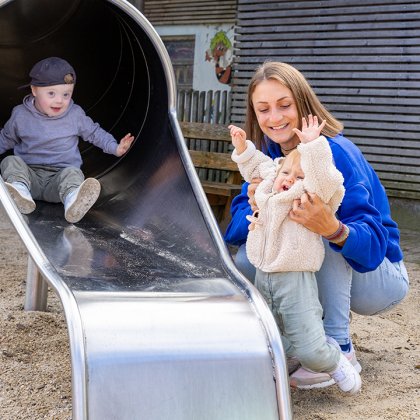 Kind rutscht eine Rutsche hinunter, Frau hält lachendes Kleinkind neben der Rutsche