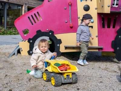 Zwei Kleinkinder spielen vor einem bunten Spielplatzgerät, eines sitzt mit einem gelben Spielzeug-LKW im Sand