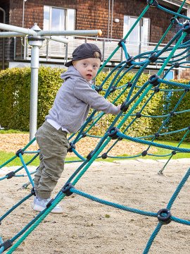 Kind klettert auf blauem Kletternetz auf Spielplatz mit Sandboden und Häusern im Hintergrund